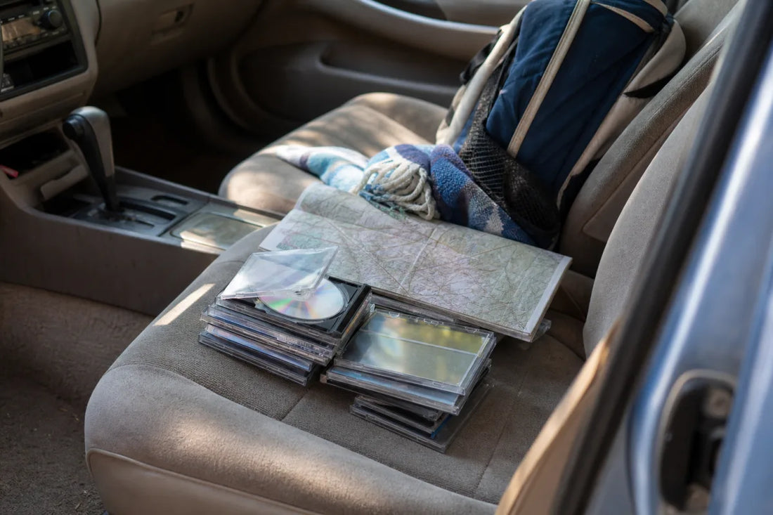 Stack of CDs in a family car with a road map and travel blanket, illustrating the best road trip CDs for families.